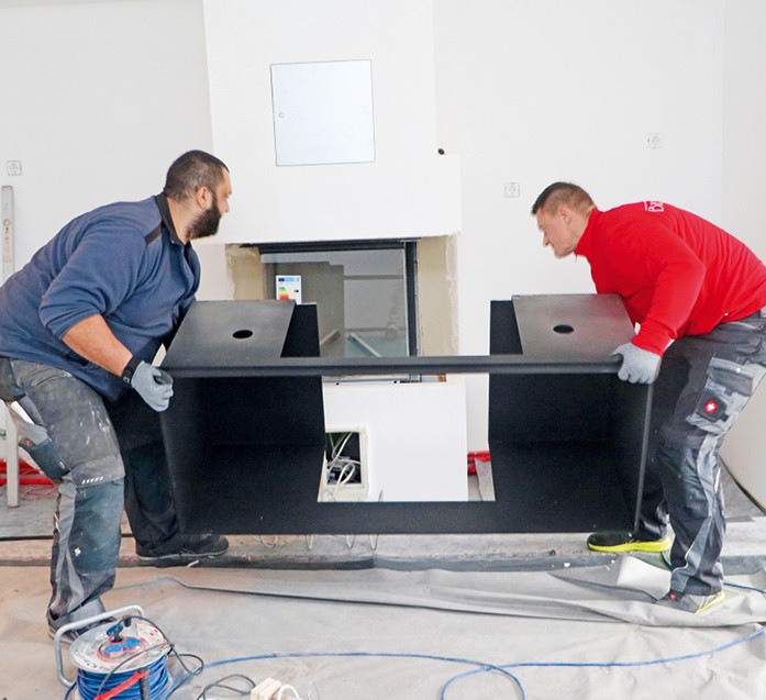 Zentrales Designelement für den Gaskamin: Ein vom Schlosser nach Architektenvorgaben maßgefertigter Rahmen aus 6 mm-Stahlblech wird von den Ofensetzern vorsichtig auf den vorbereiteten Sockel gestellt und ausgerichtet. Die beiden elektrisch beleuchtbaren Fächer links und rechts des Brennraums dienen als attraktiver Stauraum, zum Beispiel für Küchenaccessoires.