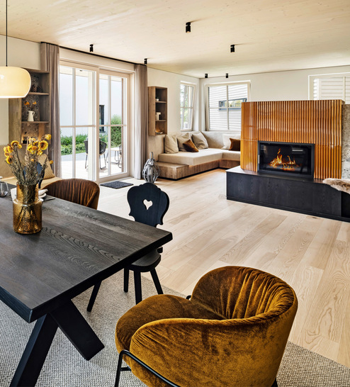 Der zentral platzierter Esstisch bietet vollen Blick auf den Ofen. Wer es noch kuscheliger möchte, kann sich die umlaufende Sitznische im Wohnzimmerbereich direkt am Kachelofen als Lieblingsplatz reservieren.